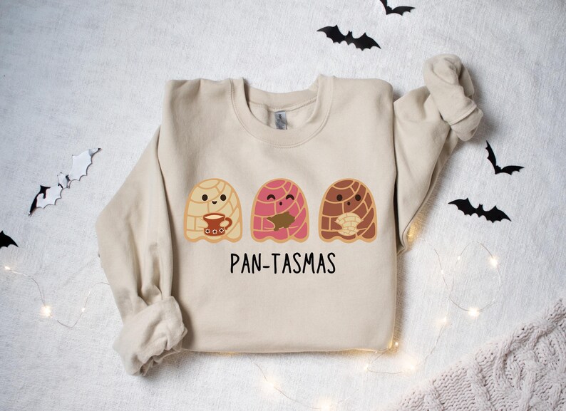 May include: A beige sweatshirt with a graphic of three cute ghosts made of pan dulce, a traditional Mexican sweet bread. The text "PAN-TASMAS" is printed below the ghosts.