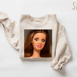 May include: Sand-colored sweatshirt featuring a square image of a doll with a surprised expression. The doll has brown hair, large eyes, and pursed lips. The word "SAND" is printed in a light brown color in the upper right corner.