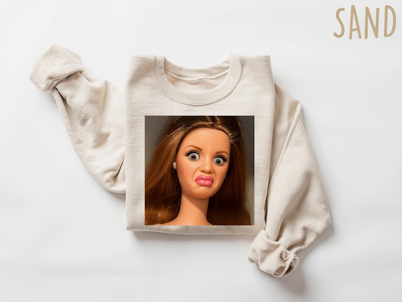 May include: Sand-colored sweatshirt featuring a square image of a doll with a surprised expression. The doll has brown hair, large eyes, and pursed lips. The word "SAND" is printed in a light brown color in the upper right corner.