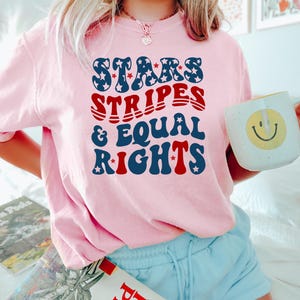 May include: A light pink sweatshirt with the text "STARS, STRIPES, & EQUAL RIGHTS" in blue and red. The sweatshirt is paired with light blue shorts. A white mug with a yellow smiley face is held in the hand.