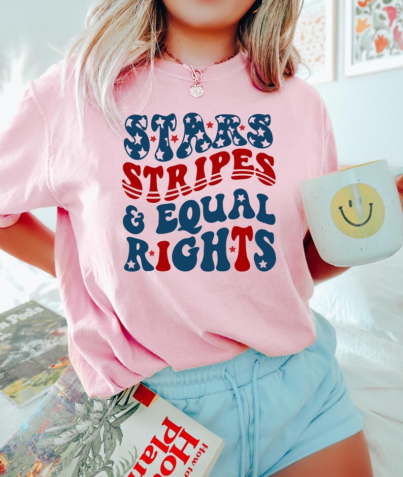 May include: A light pink sweatshirt with the text "STARS, STRIPES, & EQUAL RIGHTS" in blue and red. The sweatshirt is paired with light blue shorts. A white mug with a yellow smiley face is held in the hand.