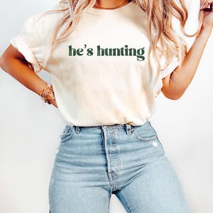 May include: Cream-colored t-shirt with the text "he's hunting" in dark green. The shirt is paired with light blue denim jeans. The overall aesthetic is casual and relaxed.