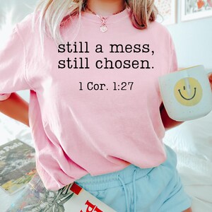 K&ouml;nnte beinhalten: Hellrosa Sweatshirt mit dem Text "still a mess, still chosen. 1 Cor. 1:27" in Schwarz. Die Person h&auml;lt einen wei&szlig;en Becher mit einem gelben Smiley. Die Person tr&auml;gt hellblaue Shorts.