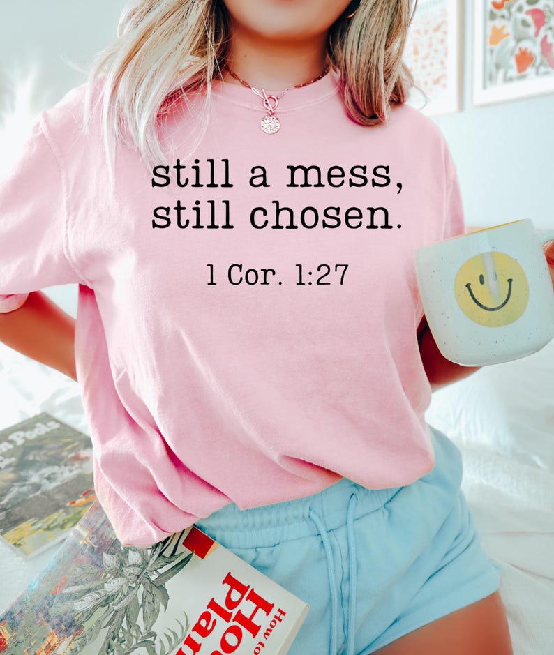 K&ouml;nnte beinhalten: Hellrosa Sweatshirt mit dem Text "still a mess, still chosen. 1 Cor. 1:27" in Schwarz. Die Person h&auml;lt einen wei&szlig;en Becher mit einem gelben Smiley. Die Person tr&auml;gt hellblaue Shorts.