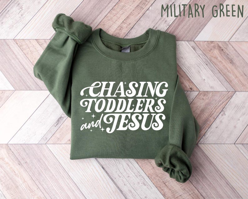 May include: Military green sweatshirt with the text "CHASING TODDLERS and JESUS" in white script. The sweatshirt is made of a soft material and has a crew neck and long sleeves. The sleeves are cuffed at the wrists.