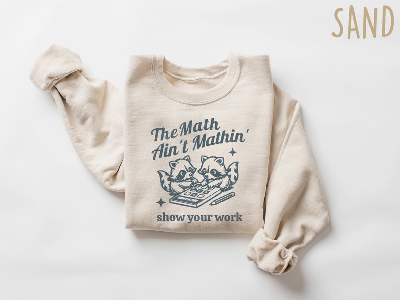 May include: A beige sweatshirt with a graphic of two raccoons using a calculator and the text "The Math Ain't Mathin' show your work". The text is in a dark blue color.