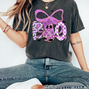 May include: Dark gray t-shirt with a Halloween-themed graphic. The design features the word "BOO" in purple and white, with a ghost and bat accents. The ghost is decorated with floral patterns and the letter "O" has a checkered pattern.
