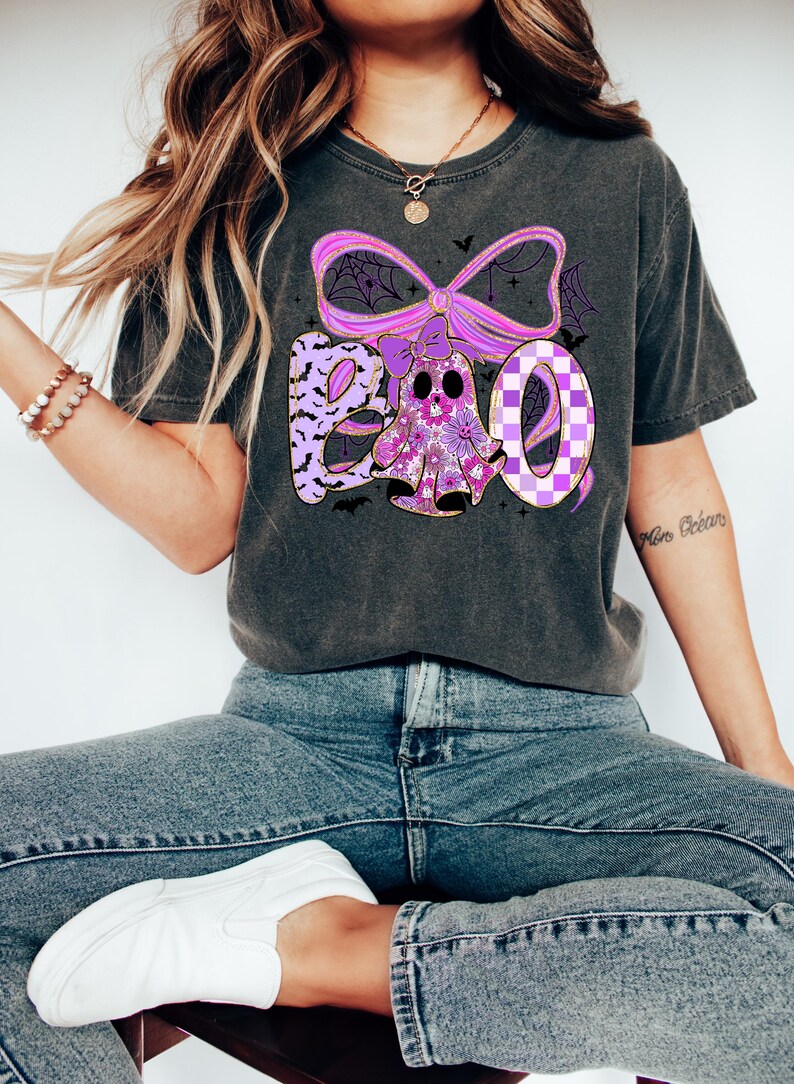 May include: Dark gray t-shirt with a Halloween-themed graphic. The design features the word "BOO" in purple and white, with a ghost and bat accents. The ghost is decorated with floral patterns and the letter "O" has a checkered pattern.