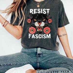May include: Dark gray t-shirt with the words "RESIST FASCISM" in white. The shirt features a black panther graphic with red flowers. The shirt is worn with blue jeans and white shoes.