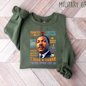 K&ouml;nnte beinhalten: Milit&auml;rgr&uuml;nes Sweatshirt mit einem Portr&auml;t von Martin Luther King Jr. und dem Text "I Have a Dream". Das Design enth&auml;lt auch die S&auml;tze "Equality for All" und "Peaceful Resistance".