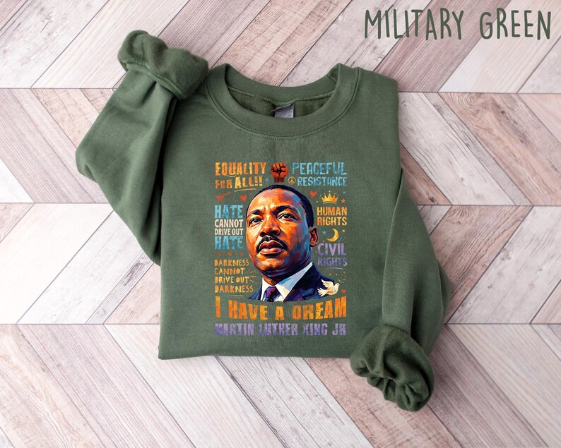 K&ouml;nnte beinhalten: Milit&auml;rgr&uuml;nes Sweatshirt mit einem Portr&auml;t von Martin Luther King Jr. und dem Text "I Have a Dream". Das Design enth&auml;lt auch die S&auml;tze "Equality for All" und "Peaceful Resistance".