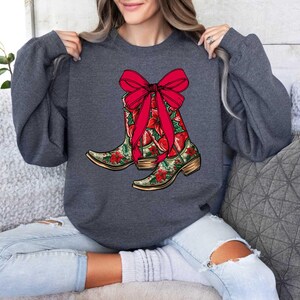 Pu&ograve; includere: Una felpa grigia scura con un design festivo di stivali da cowboy rossi e verdi con un fiocco rosso. Gli stivali sono decorati con motivi floreali.