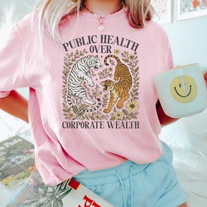 Puede incluir: Una camiseta rosa claro con el texto "PUBLIC HEALTH OVER CORPORATE WEALTH" en un dise&ntilde;o floral. El gr&aacute;fico presenta un tigre blanco y un tigre marr&oacute;n. La persona sostiene una taza blanca con una carita sonriente amarilla.