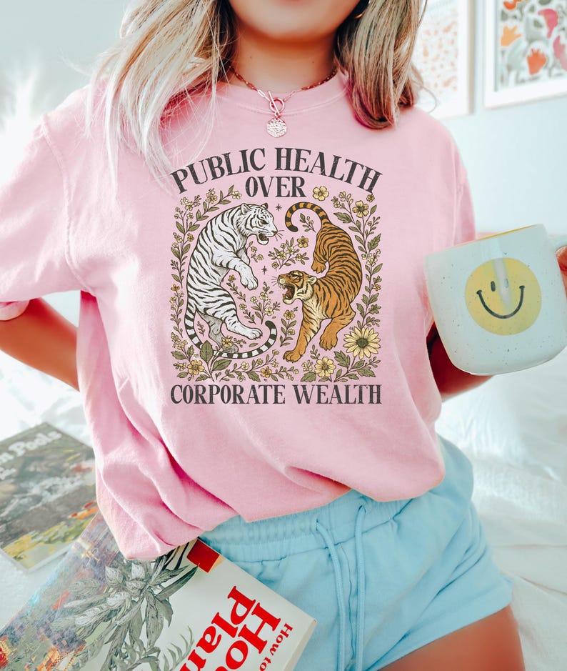 Puede incluir: Una camiseta rosa claro con el texto "PUBLIC HEALTH OVER CORPORATE WEALTH" en un dise&ntilde;o floral. El gr&aacute;fico presenta un tigre blanco y un tigre marr&oacute;n. La persona sostiene una taza blanca con una carita sonriente amarilla.