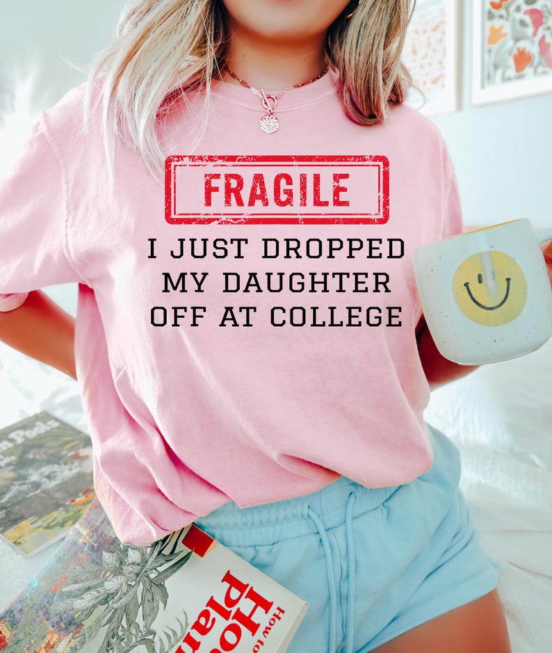 May include: A light pink sweatshirt with the text "FRAGILE I JUST DROPPED MY DAUGHTER OFF AT COLLEGE" in black and red. The person is holding a white mug with a yellow smiley face. The person is wearing light blue shorts.