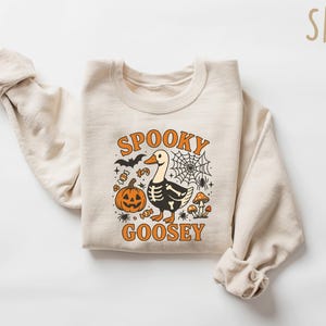 May include: A sand-colored sweatshirt with a Halloween-themed graphic. The design features a goose with a skeleton body, a jack-o'-lantern, a spiderweb, and the words "Spooky Goosey" in orange. The sweatshirt is folded.