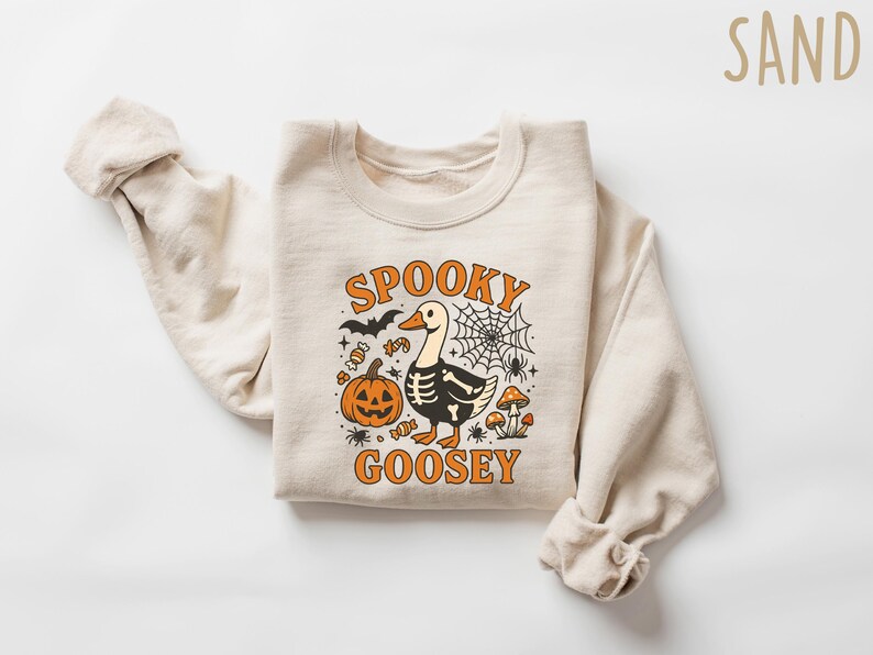 May include: A sand-colored sweatshirt with a Halloween-themed graphic. The design features a goose with a skeleton body, a jack-o'-lantern, a spiderweb, and the words "Spooky Goosey" in orange. The sweatshirt is folded.