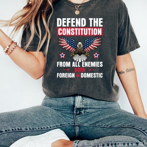 May include: Dark gray t-shirt with the text "DEFEND THE CONSTITUTION" above an eagle with American flag wings. The text "FROM ALL ENEMIES BOTH FOREIGN AND DOMESTIC" is below the eagle. The shirt is worn with blue jeans and white shoes.