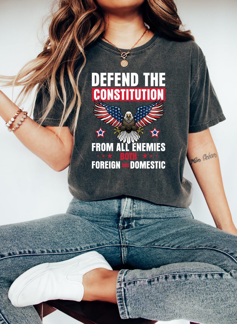 May include: Dark gray t-shirt with the text "DEFEND THE CONSTITUTION" above an eagle with American flag wings. The text "FROM ALL ENEMIES BOTH FOREIGN AND DOMESTIC" is below the eagle. The shirt is worn with blue jeans and white shoes.