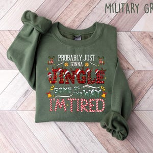 May include: A military green sweatshirt with the text "Probably Just Gonna Jingle Some of the Way I'm Tired" in red, white, and black. The word "Jingle" is in a plaid pattern. The sweatshirt is folded on a wooden surface.