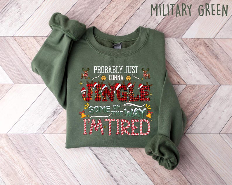 May include: A military green sweatshirt with the text "Probably Just Gonna Jingle Some of the Way I'm Tired" in red, white, and black. The word "Jingle" is in a plaid pattern. The sweatshirt is folded on a wooden surface.