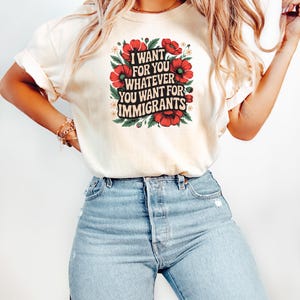May include: Cream-colored t-shirt with the text "I WANT FOR YOU WHATEVER YOU WANT FOR IMMIGRANTS" in a square design. The text is surrounded by red flowers and green leaves. The model is wearing light blue jeans.