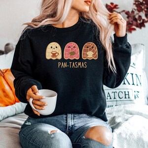 May include: Black sweatshirt with a graphic of three cartoon pan de muerto ghosts holding a cup of coffee, a book, and a pan de muerto. The text "Pan-Tasmas" is printed below the ghosts.