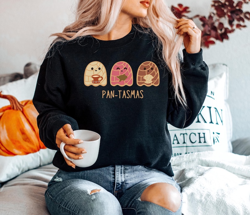 May include: Black sweatshirt with a graphic of three cartoon pan de muerto ghosts holding a cup of coffee, a book, and a pan de muerto. The text "Pan-Tasmas" is printed below the ghosts.
