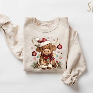May include: A cream-colored sweatshirt featuring a festive Highland cow illustration. The cow wears a Santa hat, a plaid scarf, and is adorned with Christmas lights and ornaments. The design includes snowflakes and pine branches, perfect for the holiday season.