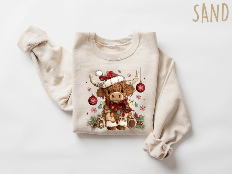 May include: A cream-colored sweatshirt featuring a festive Highland cow illustration. The cow wears a Santa hat, a plaid scarf, and is adorned with Christmas lights and ornaments. The design includes snowflakes and pine branches, perfect for the holiday season.