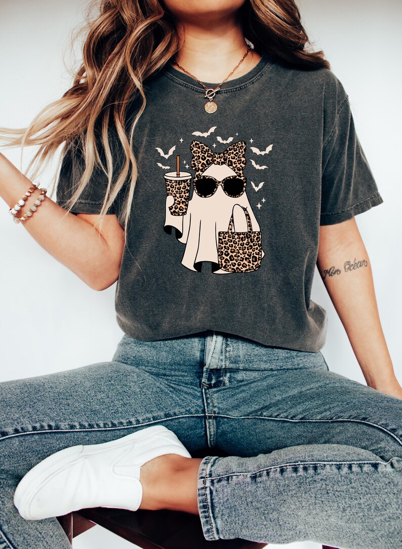 May include: Dark gray t-shirt featuring a ghost character wearing sunglasses and a leopard print bow, holding a coffee cup and a tote bag. The design includes bats and is a fun Halloween theme.