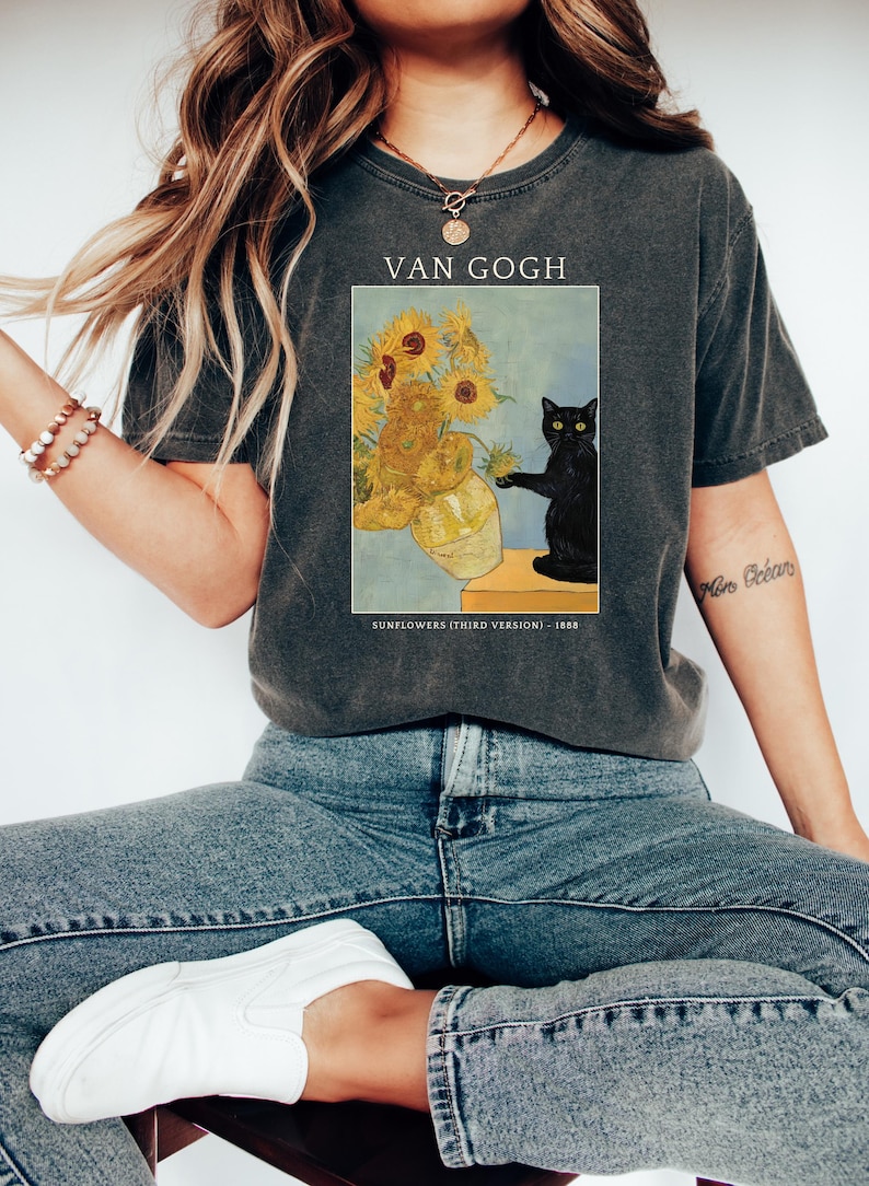 May include: Dark gray t-shirt featuring a print of Van Gogh's "Sunflowers" painting with a black cat. The text "VAN GOGH" is above the image, and "SUNFLOWERS (THIRD VERSION) - 1888" is below.
