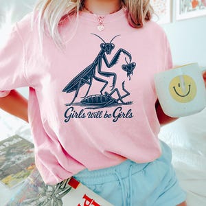 May include: Pink t-shirt with a navy blue graphic of praying mantises and the text "Girls will be Girls". The shirt is a light pink color and has a relaxed fit. The graphic features detailed insect illustrations.