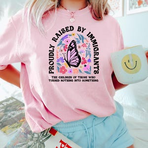 May include: Pink sweatshirt with the text "PROUDLY RAISED BY IMMIGRANTS" in a curved arch design. A butterfly graphic is centered within the arch, surrounded by colorful flowers. The text "THE CHILDREN OF THOSE WHO TURNED NOTHING INTO SOMETHING" is below.