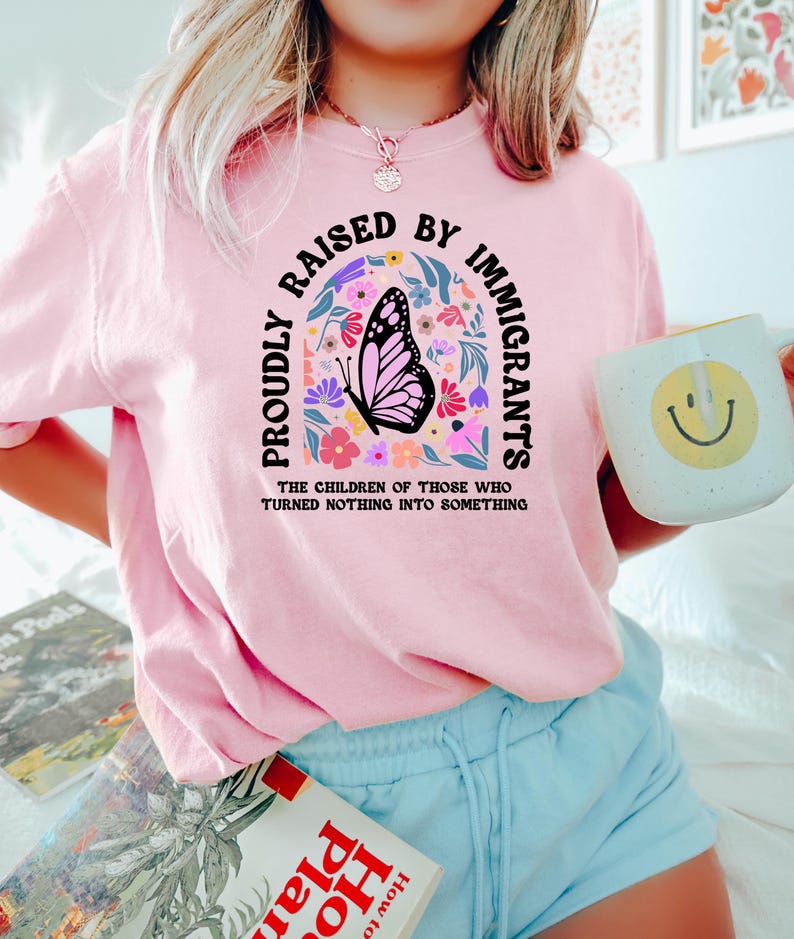 May include: Pink sweatshirt with the text "PROUDLY RAISED BY IMMIGRANTS" in a curved arch design. A butterfly graphic is centered within the arch, surrounded by colorful flowers. The text "THE CHILDREN OF THOSE WHO TURNED NOTHING INTO SOMETHING" is below.