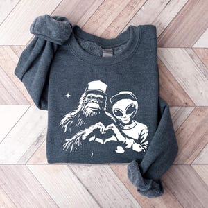 Puede incluir: Una sudadera gris oscura con un estampado blanco de un Bigfoot y un extraterrestre haciendo un coraz&oacute;n con sus manos. El extraterrestre lleva una gorra de b&eacute;isbol.