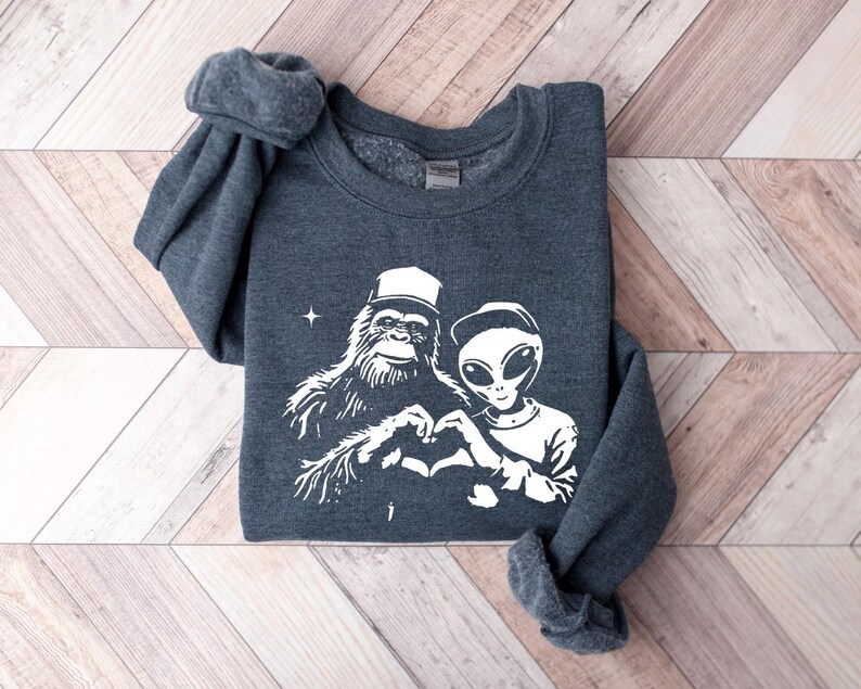 Puede incluir: Una sudadera gris oscura con un estampado blanco de un Bigfoot y un extraterrestre haciendo un coraz&oacute;n con sus manos. El extraterrestre lleva una gorra de b&eacute;isbol.