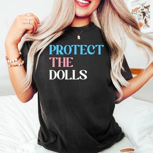 May include: Black t-shirt with the text "PROTECT THE DOLLS" in blue and pink letters.