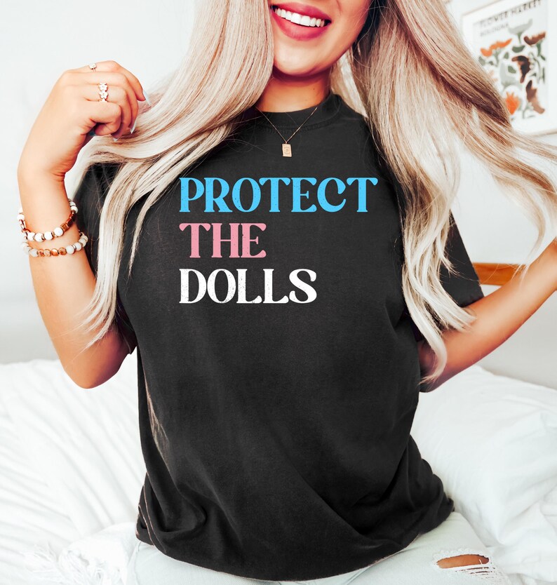 May include: Black t-shirt with the text "PROTECT THE DOLLS" in blue and pink letters.