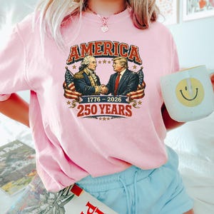 Puede incluir: Camiseta rosa claro con un gr&aacute;fico que dice "AMERICA" sobre una imagen de dos hombres d&aacute;ndose la mano, con banderas americanas a cada lado. Tambi&eacute;n se muestra el texto "1776-2026" y "250 YEARS". Se sostiene una taza blanca con una carita sonriente.
