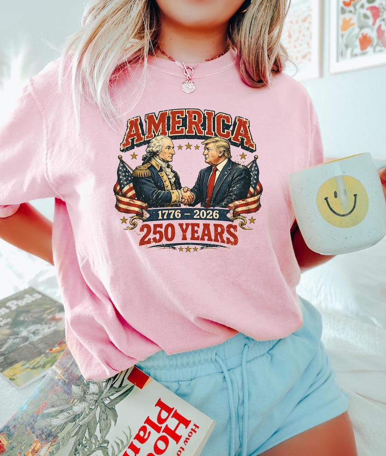 Puede incluir: Camiseta rosa claro con un gr&aacute;fico que dice "AMERICA" sobre una imagen de dos hombres d&aacute;ndose la mano, con banderas americanas a cada lado. Tambi&eacute;n se muestra el texto "1776-2026" y "250 YEARS". Se sostiene una taza blanca con una carita sonriente.