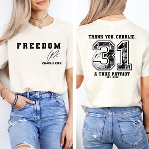 May include: Cream-colored t-shirt with the word "FREEDOM" in bold black letters on the front, along with a signature and the name "CHARLIE KIRK." The back of the shirt features the text "THANK YOU, CHARLIE," the number "31," and the words "A TRUE PATRIOT 1993-2020."