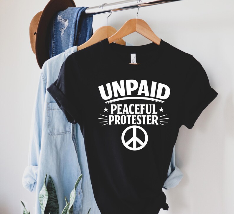 May include: Black t-shirt with the white text "UNPAID PEACEFUL PROTESTER" and a peace symbol. The shirt is hanging on a wooden hanger with a denim shirt and a brown hat in the background.
