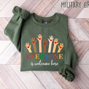 May include: Military green sweatshirt with the text "Everyone is welcome here." The design features hands of various skin tones, each with a red heart. The sweatshirt is made of a soft material and is perfect for casual wear.