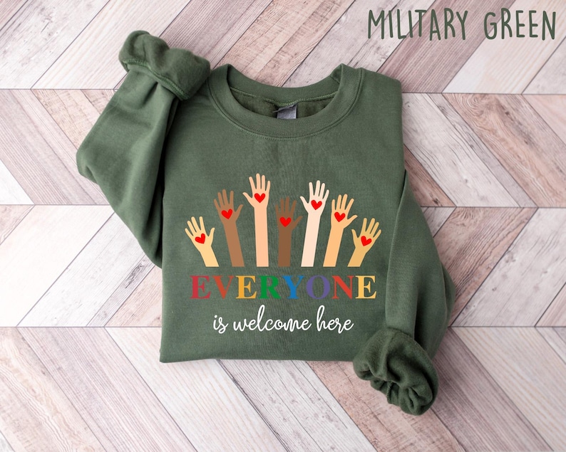 May include: Military green sweatshirt with the text "Everyone is welcome here." The design features hands of various skin tones, each with a red heart. The sweatshirt is made of a soft material and is perfect for casual wear.