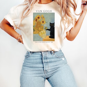 May include: Cream-colored t-shirt featuring a print of Van Gogh's "Sunflowers (Third Version)" painting, with a black cat. The text "VAN GOGH" is above the artwork, and "SUNFLOWERS (THIRD VERSION) - 1888" is below. The shirt is paired with blue jeans.