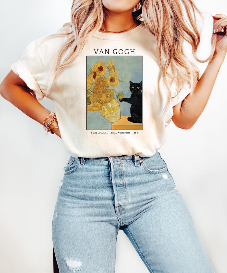 May include: Cream-colored t-shirt featuring a print of Van Gogh's "Sunflowers (Third Version)" painting, with a black cat. The text "VAN GOGH" is above the artwork, and "SUNFLOWERS (THIRD VERSION) - 1888" is below. The shirt is paired with blue jeans.