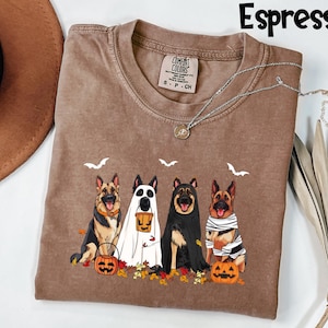 May include: Espresso-colored t-shirt featuring a Halloween-themed graphic of four German Shepherds in costumes. The dogs are dressed as a ghost, a mummy, and a vampire, with pumpkins and fall leaves. The shirt has a Comfort Colors tag.