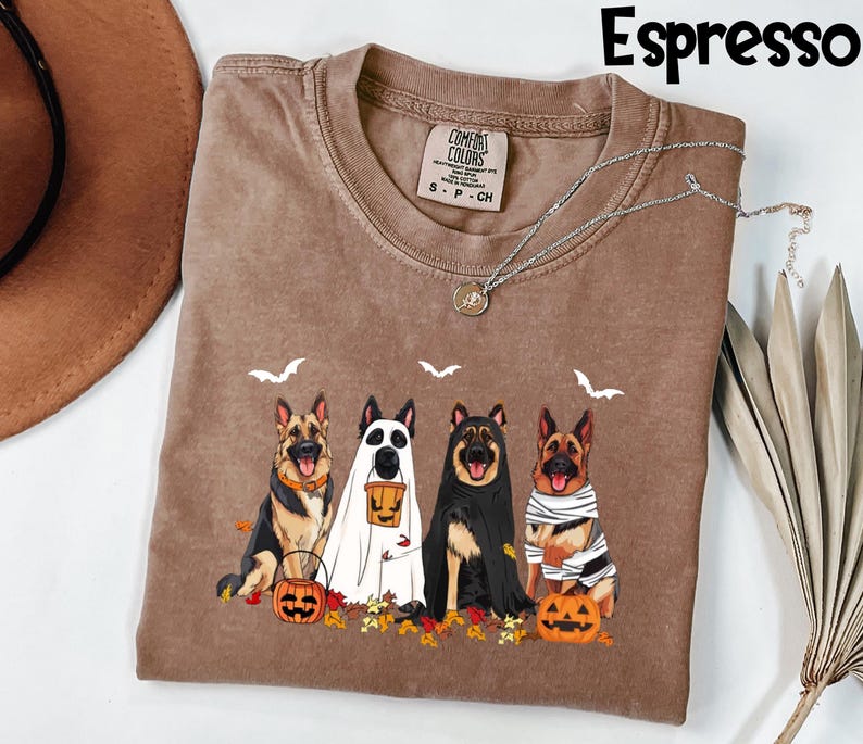 May include: Espresso-colored t-shirt featuring a Halloween-themed graphic of four German Shepherds in costumes. The dogs are dressed as a ghost, a mummy, and a vampire, with pumpkins and fall leaves. The shirt has a Comfort Colors tag.