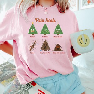 Puede incluir: Camiseta rosa con el texto "Pain Scale" e ilustraciones de &aacute;rboles de Navidad que representan niveles de dolor. La camiseta tambi&eacute;n presenta los textos "No Pain", "Mild Pain", "Moderate Pain", "Severe", "Very Severe Pain" y "Worst Pain Possible".